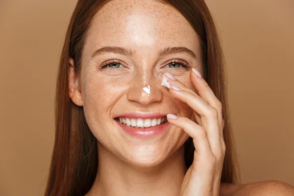 Uśmiechnięta kobieta z cerą pokrytą piegami nakłada krem na twarz.